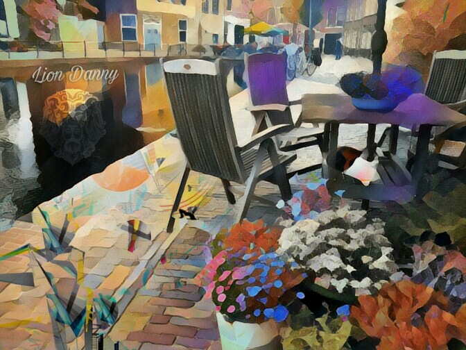 Abstract art of a waterway to the left an a street to the right, populated by a table wit chairs and a parasol, near bunches of colorful flowers, a cat is under the table